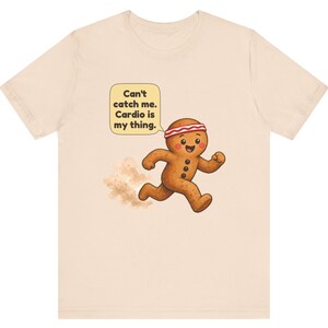 May include: A light beige t-shirt featuring a graphic of a running gingerbread person wearing a red and white headband. A speech bubble above the character contains the text: Can't catch me. Cardio is my thing. A small dust cloud appears behind the figure.