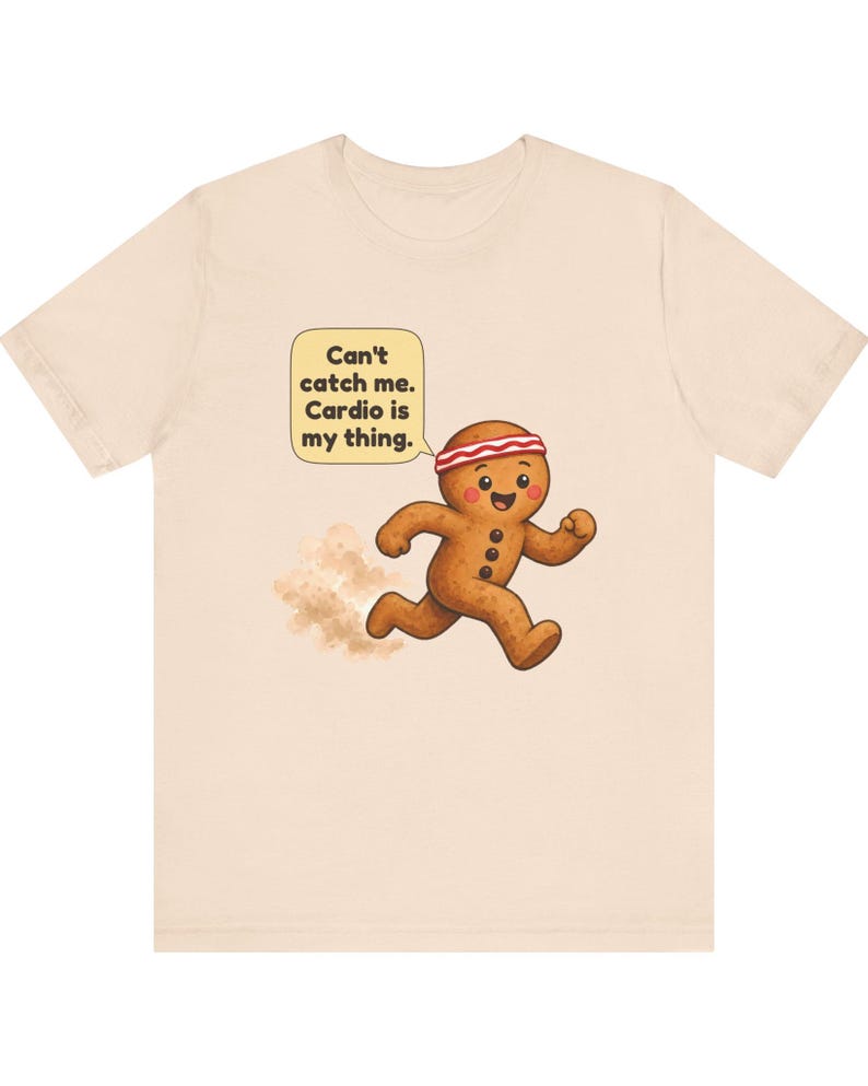 May include: A light beige t-shirt featuring a graphic of a running gingerbread person wearing a red and white headband. A speech bubble above the character contains the text: Can't catch me. Cardio is my thing. A small dust cloud appears behind the figure.