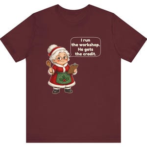 K&ouml;nnte beinhalten: Ein bordeauxrotes Kurzarm-T-Shirt mit einer Cartoon-Illustration einer &auml;lteren Frau in einem rot-wei&szlig;en Weihnachtsmann-Outfit, die einen Holzl&ouml;ffel und ein Klemmbrett h&auml;lt, mit einer Sprechblase: I run the workshop. He gets the credit.