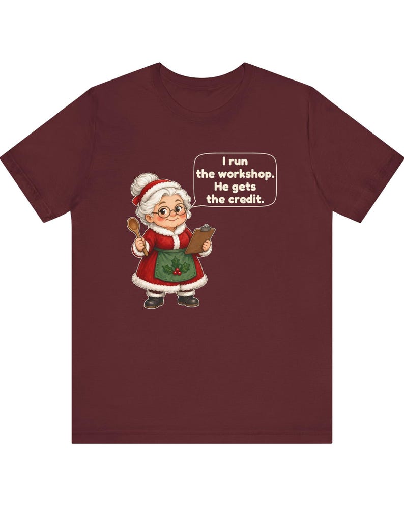 K&ouml;nnte beinhalten: Ein bordeauxrotes Kurzarm-T-Shirt mit einer Cartoon-Illustration einer &auml;lteren Frau in einem rot-wei&szlig;en Weihnachtsmann-Outfit, die einen Holzl&ouml;ffel und ein Klemmbrett h&auml;lt, mit einer Sprechblase: I run the workshop. He gets the credit.