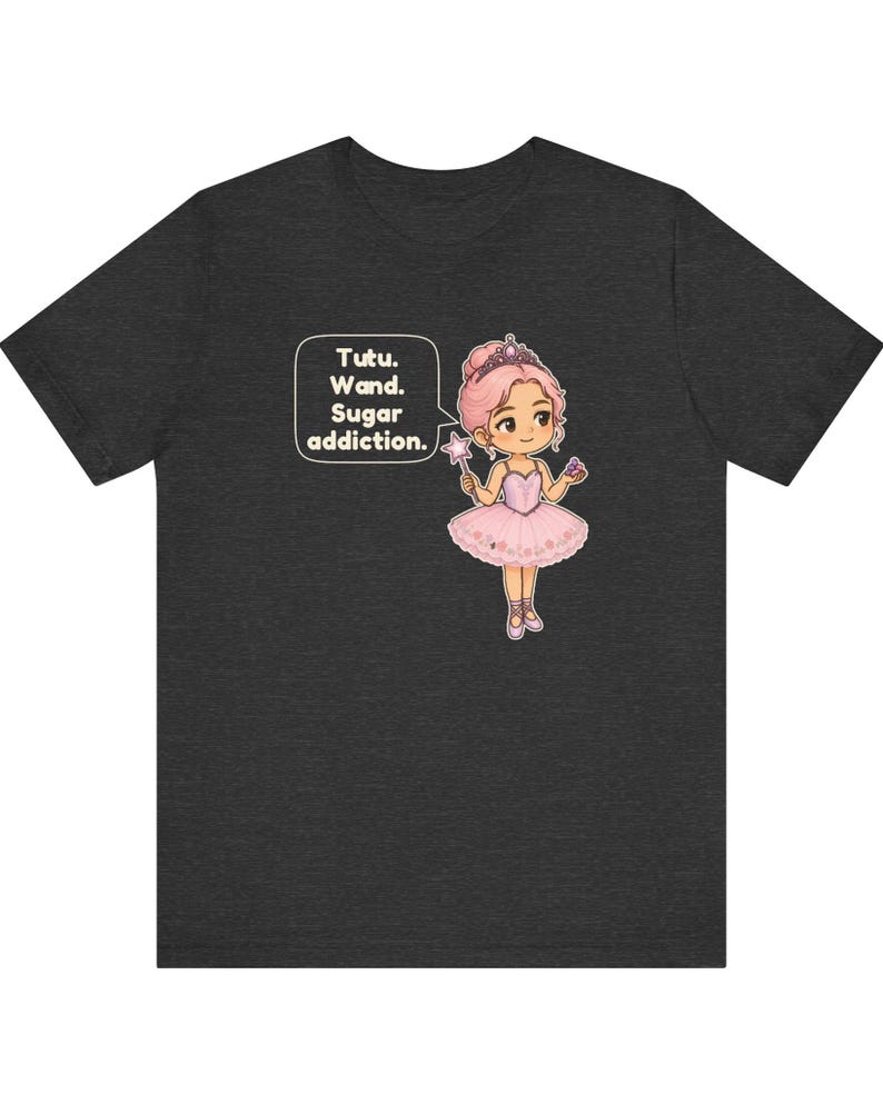 May include: A dark heather gray t-shirt featuring a cartoon illustration of a ballerina with pink hair, a tiara, and a pink tutu. The character holds a star-tipped wand and a cupcake, with a speech bubble that reads: Tutu. Wand. Sugar addiction.
