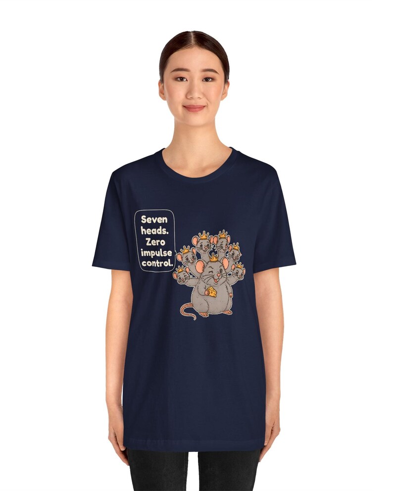 May include: A navy blue t-shirt featuring a graphic of a seven-headed rat wearing small gold crowns and holding a piece of yellow cheese. A speech bubble next to the rat contains the text: Seven heads. Zero impulse control.