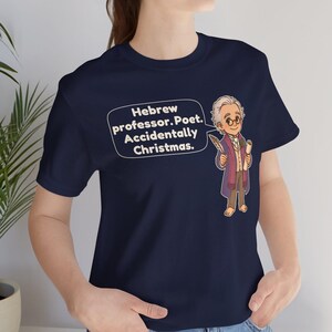 Pu&ograve; includere: Una t-shirt blu navy a maniche corte con un'illustrazione a cartoni animati di un uomo anziano con gli occhiali che tiene in mano una penna e una pergamena. Un fumetto contiene il testo: Hebrew professor. Poet. Accidentally Christmas.