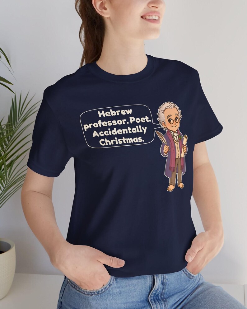 Pu&ograve; includere: Una t-shirt blu navy a maniche corte con un'illustrazione a cartoni animati di un uomo anziano con gli occhiali che tiene in mano una penna e una pergamena. Un fumetto contiene il testo: Hebrew professor. Poet. Accidentally Christmas.
