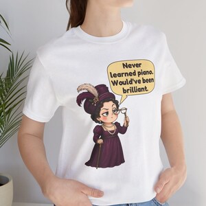May include: A white t-shirt featuring a graphic of a person in a purple Victorian-style dress, a feathered hat, and pearls, holding a monocle. A yellow speech bubble contains the text: Never learned piano. Would've been brilliant.