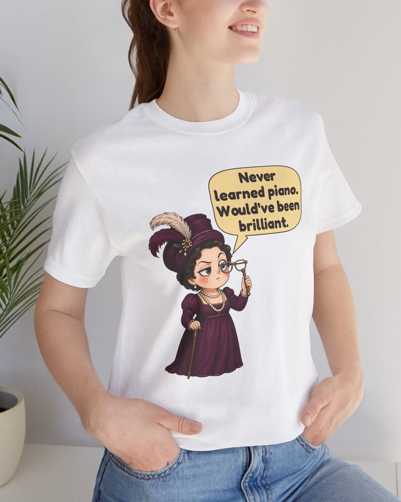 May include: A white t-shirt featuring a graphic of a person in a purple Victorian-style dress, a feathered hat, and pearls, holding a monocle. A yellow speech bubble contains the text: Never learned piano. Would've been brilliant.