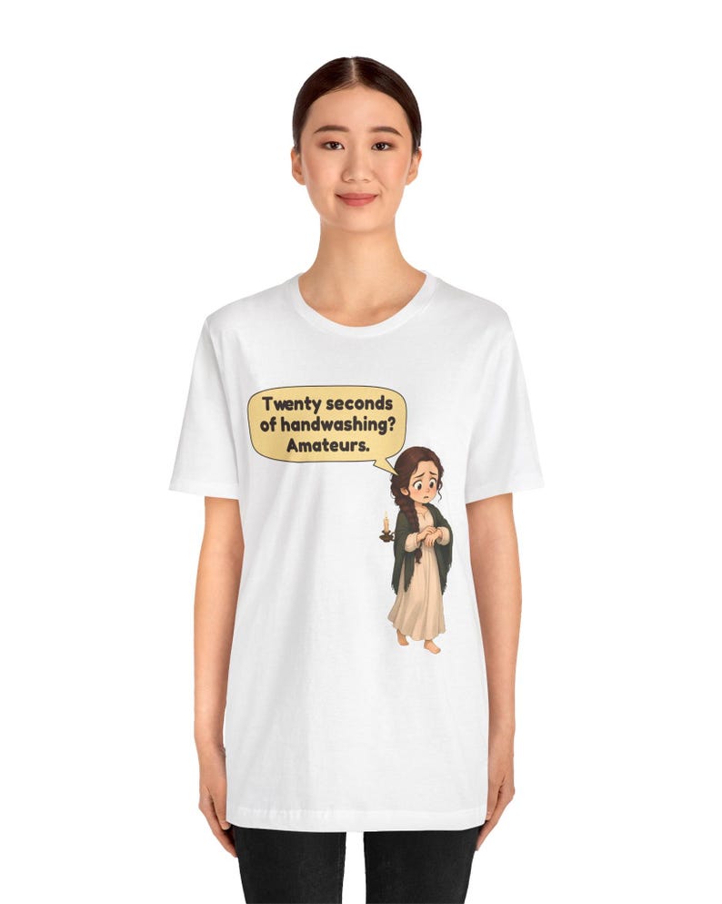 Op de afbeelding: Een wit t-shirt met een cartoonillustratie van een persoon met bruin haar in een lange jurk die een kaars vasthoudt. Een tekstballon boven het personage bevat de tekst: Twenty seconds of handwashing? Amateurs.