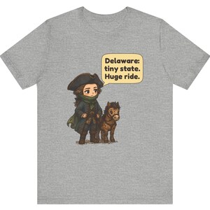 May include: A heather gray t-shirt featuring a cartoon illustration of a person in a tricorn hat and colonial-style clothing standing next to a small brown horse. A speech bubble contains the text: Delaware: tiny state. Huge ride.