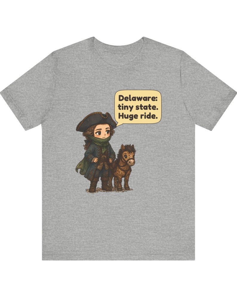 May include: A heather gray t-shirt featuring a cartoon illustration of a person in a tricorn hat and colonial-style clothing standing next to a small brown horse. A speech bubble contains the text: Delaware: tiny state. Huge ride.