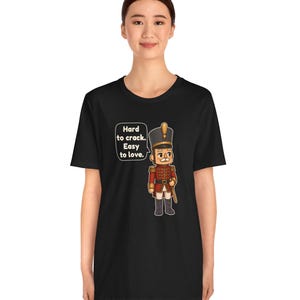 Pu&ograve; includere: Una t-shirt nera con la grafica di un soldatino schiaccianoci in uniforme rossa con un cappello alto. Accanto al soldatino c'&egrave; un fumetto con il testo: Hard to crack. Easy to love. Il disegno &egrave; centrato sulla parte anteriore della maglietta.