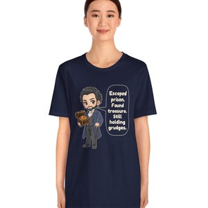 May include: A navy blue t-shirt featuring a cartoon illustration of a man in a dark coat holding a treasure chest, with a speech bubble containing the text: Escaped prison. Found treasure. Still holding grudges.