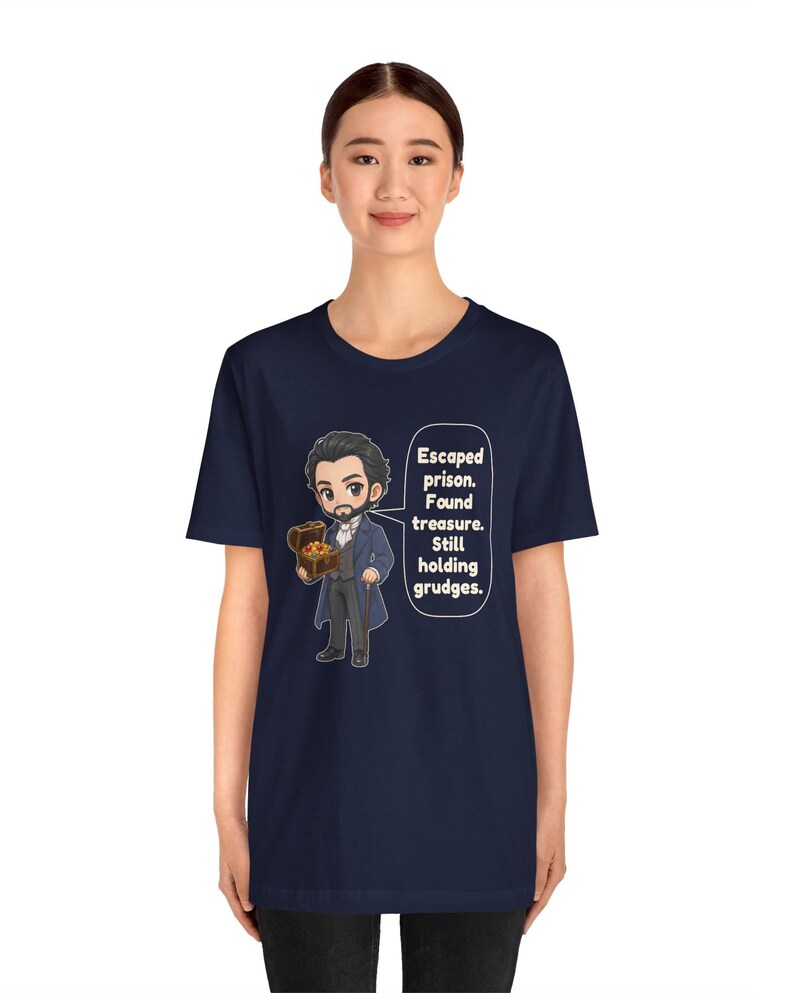 May include: A navy blue t-shirt featuring a cartoon illustration of a man in a dark coat holding a treasure chest, with a speech bubble containing the text: Escaped prison. Found treasure. Still holding grudges.