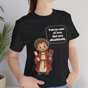 Puede incluir: Una camiseta negra con una ilustraci&oacute;n de dibujos animados de una figura barbuda con t&uacute;nica roja que sostiene un pergamino y una rosa roja. Un globo de texto sobre la figura contiene el texto: Patron saint of love. Not your situationship.