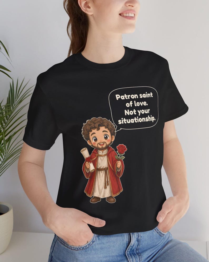 Puede incluir: Una camiseta negra con una ilustraci&oacute;n de dibujos animados de una figura barbuda con t&uacute;nica roja que sostiene un pergamino y una rosa roja. Un globo de texto sobre la figura contiene el texto: Patron saint of love. Not your situationship.