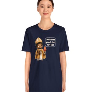 May include: A navy blue t-shirt featuring a cartoon illustration of a bishop in gold vestments holding a book and a flaming heart, with a speech bubble containing the text: Make me good. Just not yet.