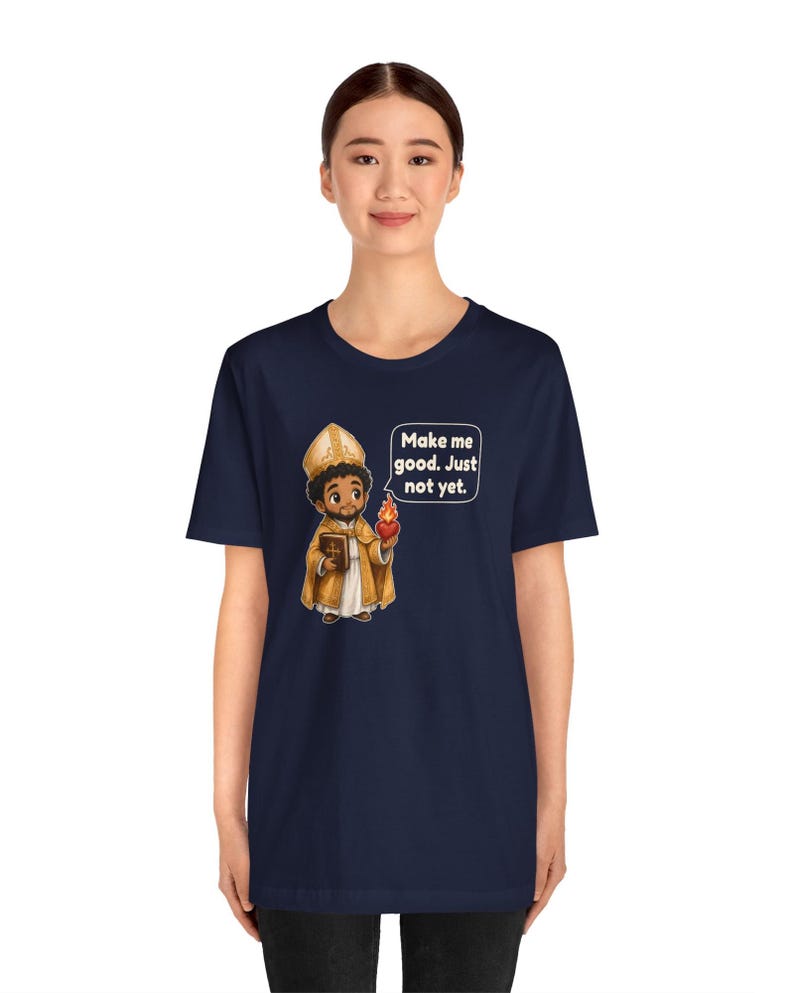May include: A navy blue t-shirt featuring a cartoon illustration of a bishop in gold vestments holding a book and a flaming heart, with a speech bubble containing the text: Make me good. Just not yet.