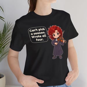 May include: A black t-shirt featuring a cartoon illustration of a red-haired violinist in a dark robe holding a violin. A speech bubble contains the text: Can't pick a season. Wrote all four. The design is centered on the front of the garment.