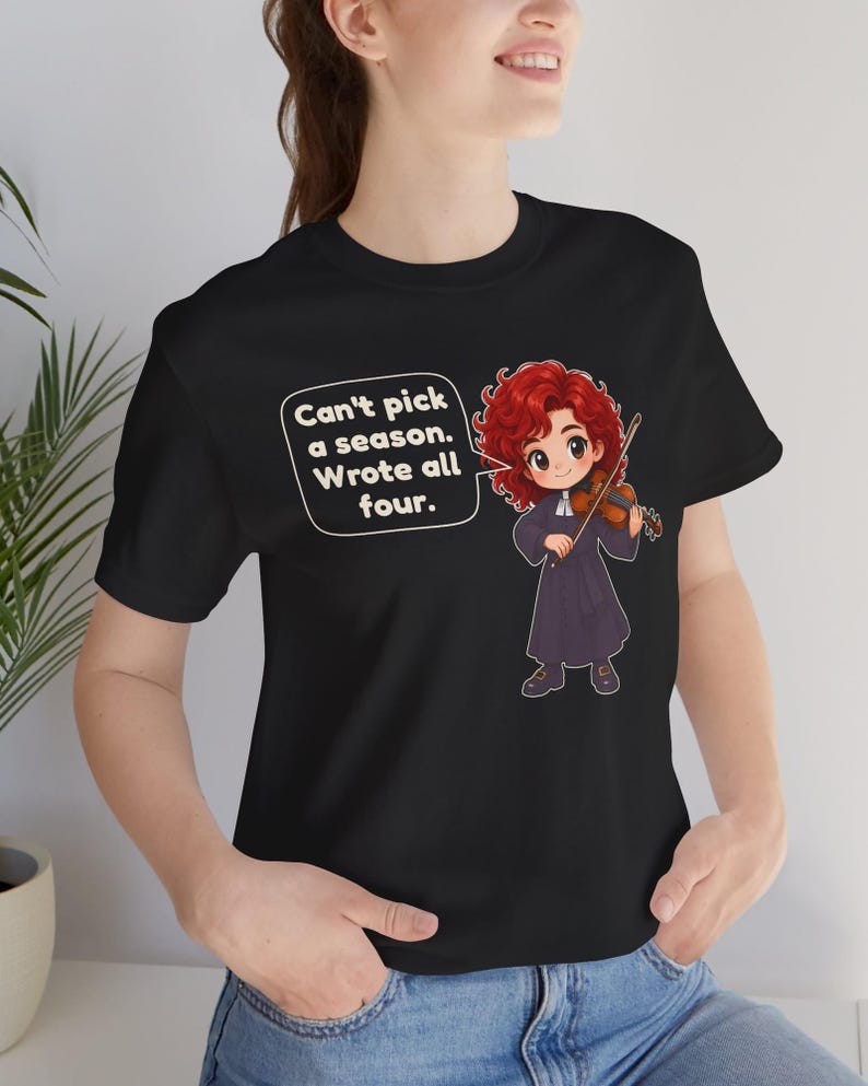 May include: A black t-shirt featuring a cartoon illustration of a red-haired violinist in a dark robe holding a violin. A speech bubble contains the text: Can't pick a season. Wrote all four. The design is centered on the front of the garment.