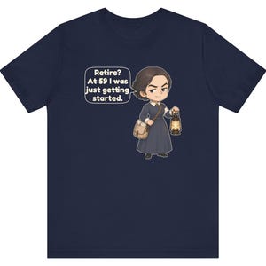 May include: A navy blue short-sleeve t-shirt featuring a cartoon illustration of a person in a dark dress carrying a satchel and a lit lantern. A speech bubble above the figure contains the text: Retire? At 59 I was just getting started.