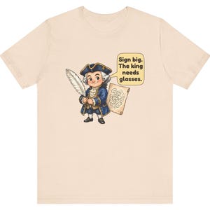 May include: A light beige t-shirt featuring a cartoon illustration of a person in an 18th-century blue coat and tricorn hat holding a white quill and parchment. A speech bubble contains the text: Sign big. The king needs glasses.
