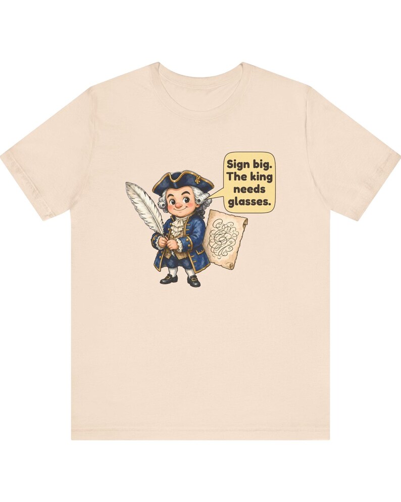 May include: A light beige t-shirt featuring a cartoon illustration of a person in an 18th-century blue coat and tricorn hat holding a white quill and parchment. A speech bubble contains the text: Sign big. The king needs glasses.