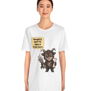 Puede incluir: Una camiseta blanca con un gr&aacute;fico de una peque&ntilde;a criatura marr&oacute;n con cuernos que sostiene un manojo de palos y un pergamino. Un globo de texto amarillo sobre la criatura contiene el texto: Naughty list? I'll take it from here.