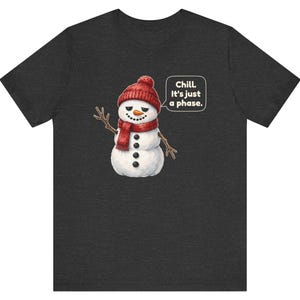 Pode incluir: Uma t-shirt cinzento-escura de manga curta com o gr&aacute;fico de um boneco de neve a usar um gorro e um cachecol de malha vermelhos. O boneco de neve tem um nariz de cenoura, bra&ccedil;os de ramo e um bal&atilde;o com o texto, Chill. It's just a phase.