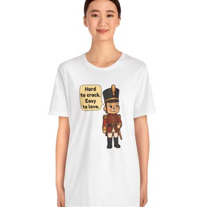 Pu&ograve; includere: Una t-shirt bianca a maniche corte con la grafica di un soldatino schiaccianoci in stile cartoon, con uniforme rossa e cappello nero. Accanto al personaggio c'&egrave; un fumetto con il testo: Hard to crack. Easy to love.