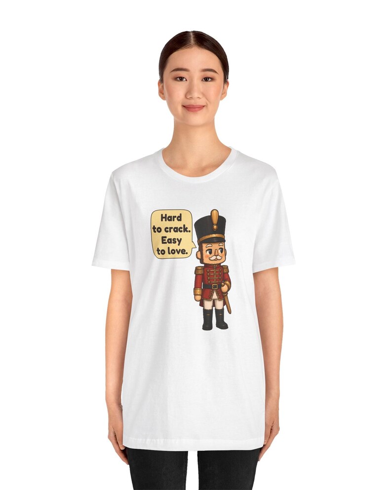 Pu&ograve; includere: Una t-shirt bianca a maniche corte con la grafica di un soldatino schiaccianoci in stile cartoon, con uniforme rossa e cappello nero. Accanto al personaggio c'&egrave; un fumetto con il testo: Hard to crack. Easy to love.