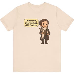 May include: A light beige t-shirt featuring a cartoon illustration of a person in a brown coat and white scarf holding a quill and candle. A speech bubble next to the character contains the text: Underpaid, overworked, still festive.