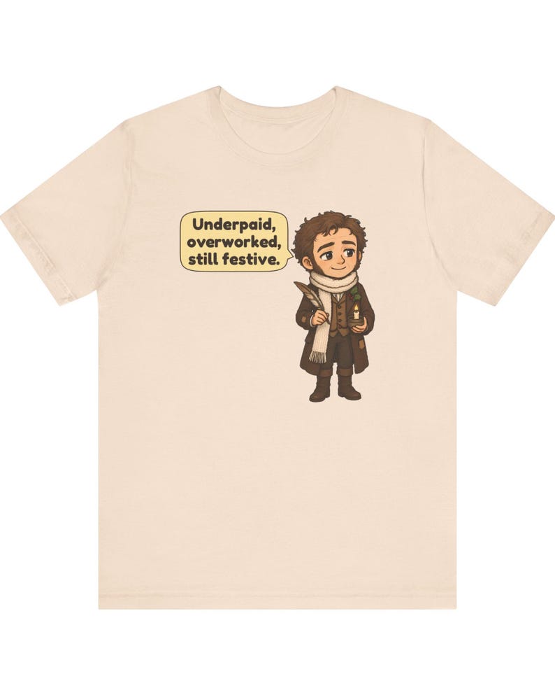 May include: A light beige t-shirt featuring a cartoon illustration of a person in a brown coat and white scarf holding a quill and candle. A speech bubble next to the character contains the text: Underpaid, overworked, still festive.