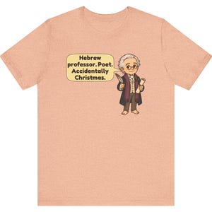 Pu&ograve; includere: Una t-shirt a maniche corte color pesca m&eacute;lange con un'illustrazione a cartone animato di un personaggio che tiene una penna e una pergamena. Un fumetto contiene il testo: Hebrew professor. Poet. Accidentally Christmas.