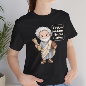 May include: A black t-shirt featuring a cartoon illustration of a white-haired, bearded figure in a light blue and beige tunic holding a scroll and a brown jug, with a speech bubble containing the text: First, do no harm. Second, coffee.