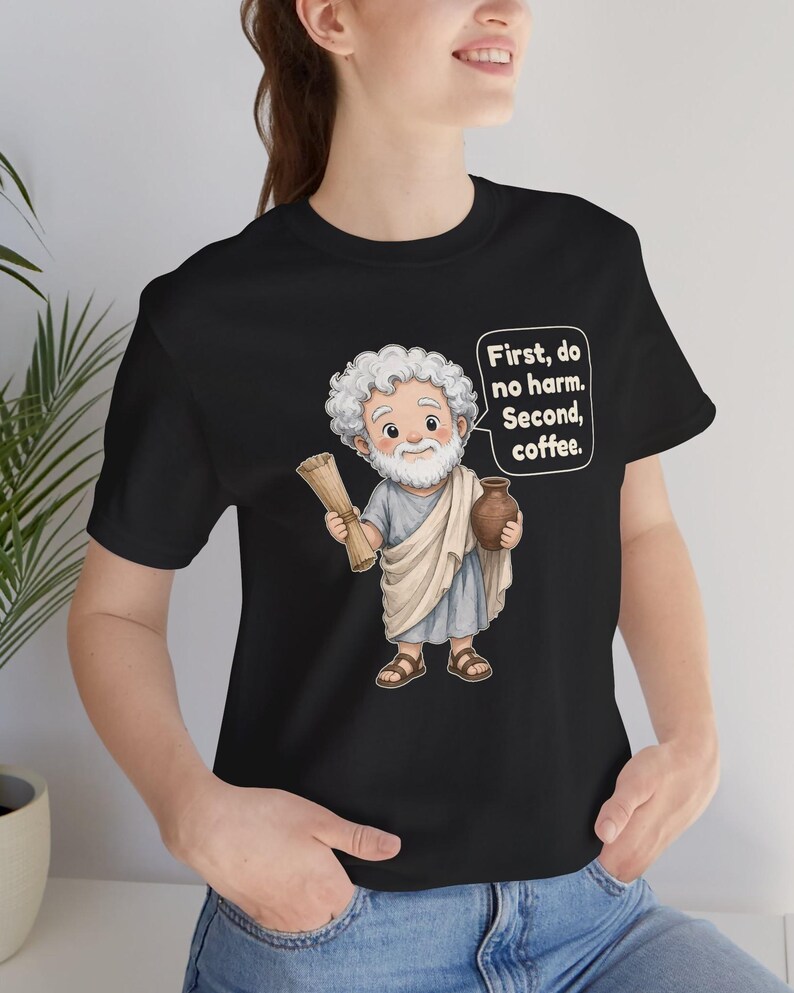 May include: A black t-shirt featuring a cartoon illustration of a white-haired, bearded figure in a light blue and beige tunic holding a scroll and a brown jug, with a speech bubble containing the text: First, do no harm. Second, coffee.