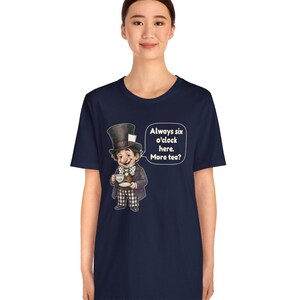 May include: A navy blue t-shirt featuring a graphic of a character wearing a tall top hat, a polka dot bow tie, and a suit, holding a teacup and saucer. Next to the character is a speech bubble with the text: Always six o'clock here. More tea?
