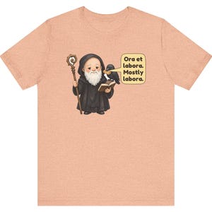 May include: A peach-colored t-shirt featuring a graphic of a monk in a black robe holding a wooden staff and an open book. A black raven sits on the monk's shoulder. A speech bubble contains the text: Ora et labora. Mostly labora.
