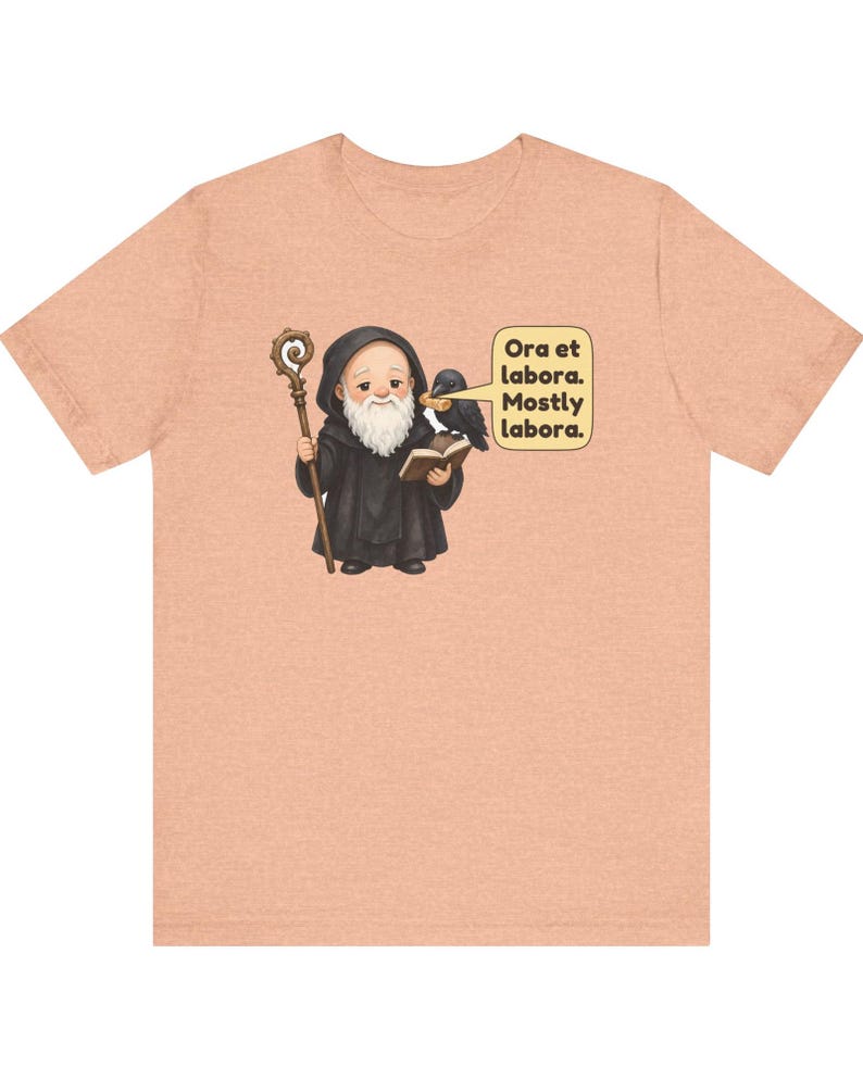 May include: A peach-colored t-shirt featuring a graphic of a monk in a black robe holding a wooden staff and an open book. A black raven sits on the monk's shoulder. A speech bubble contains the text: Ora et labora. Mostly labora.