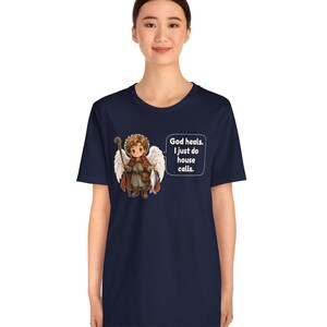 May include: A navy blue t-shirt featuring a graphic of a cartoon angel with brown hair, white wings, a red cloak, and a wooden staff. The angel holds a fish, and a speech bubble contains the text: God heals. I just do house calls.