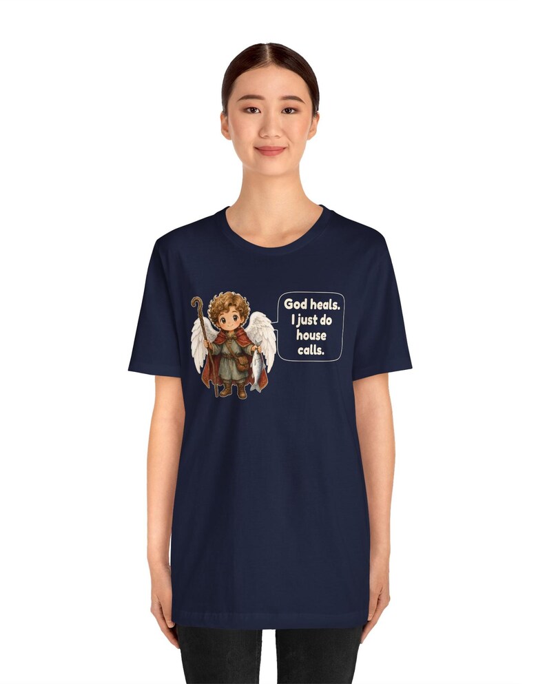 May include: A navy blue t-shirt featuring a graphic of a cartoon angel with brown hair, white wings, a red cloak, and a wooden staff. The angel holds a fish, and a speech bubble contains the text: God heals. I just do house calls.