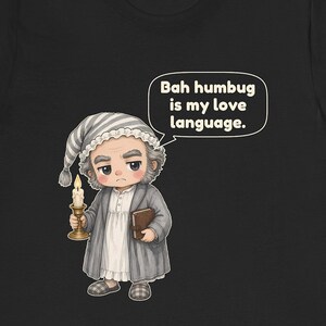 Puede incluir: Una camiseta negra con una ilustraci&oacute;n de dibujos animados de un personaje con gorro de dormir y bata a rayas grises, sosteniendo una vela encendida y un libro. Un bocadillo contiene el texto: Bah humbug is my love language.