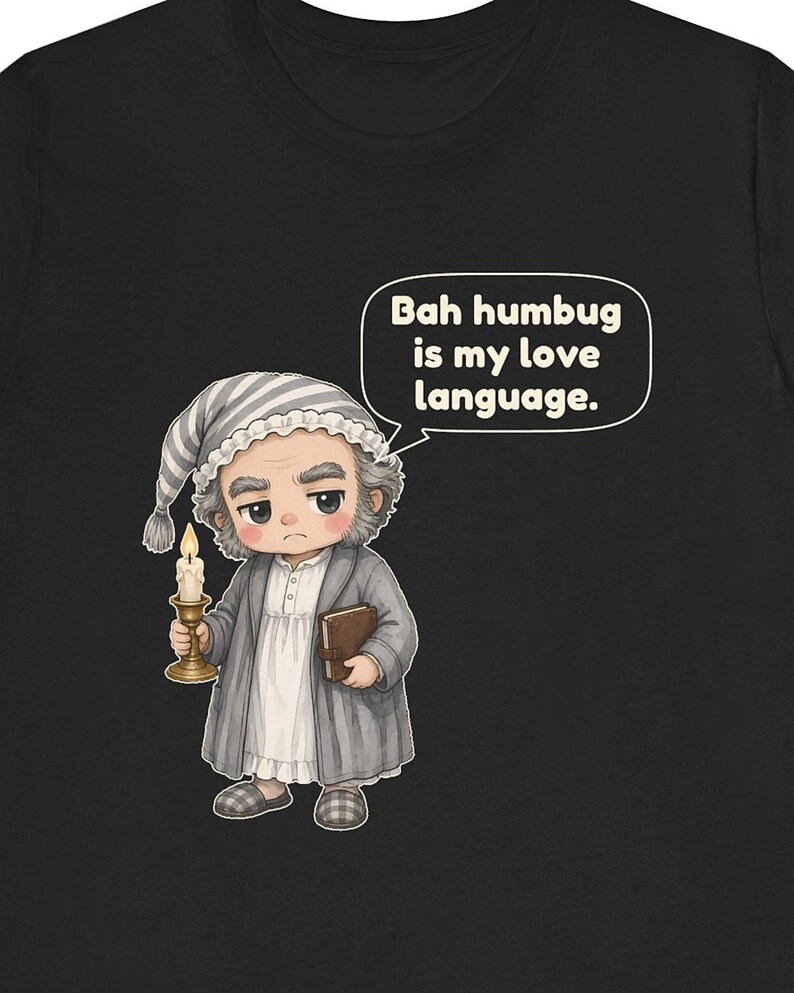 Puede incluir: Una camiseta negra con una ilustraci&oacute;n de dibujos animados de un personaje con gorro de dormir y bata a rayas grises, sosteniendo una vela encendida y un libro. Un bocadillo contiene el texto: Bah humbug is my love language.