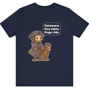 May include: A navy blue t-shirt featuring a graphic of a person in a historical tricorn hat and purple coat standing next to a small brown horse. A speech bubble above them contains the text: Delaware: tiny state. Huge ride.