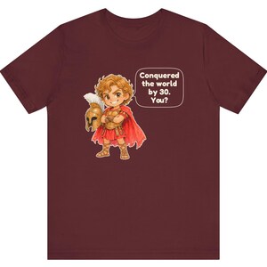 May include: A maroon short-sleeve t-shirt featuring a cartoon illustration of a warrior in golden armor and a red cape holding a helmet. A speech bubble next to the character contains the text: Conquered the world by 30. You?