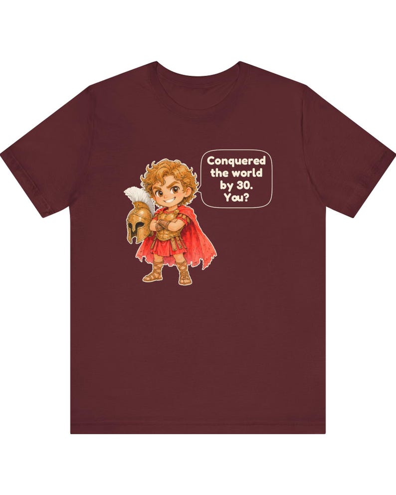 May include: A maroon short-sleeve t-shirt featuring a cartoon illustration of a warrior in golden armor and a red cape holding a helmet. A speech bubble next to the character contains the text: Conquered the world by 30. You?