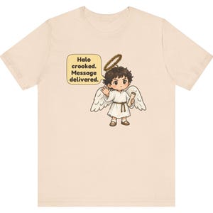 May include: A light beige t-shirt featuring a graphic of a cartoon angel with dark hair, white wings, and a crooked halo. The angel holds a scroll and waves, with a speech bubble containing the text: Halo crooked. Message delivered.