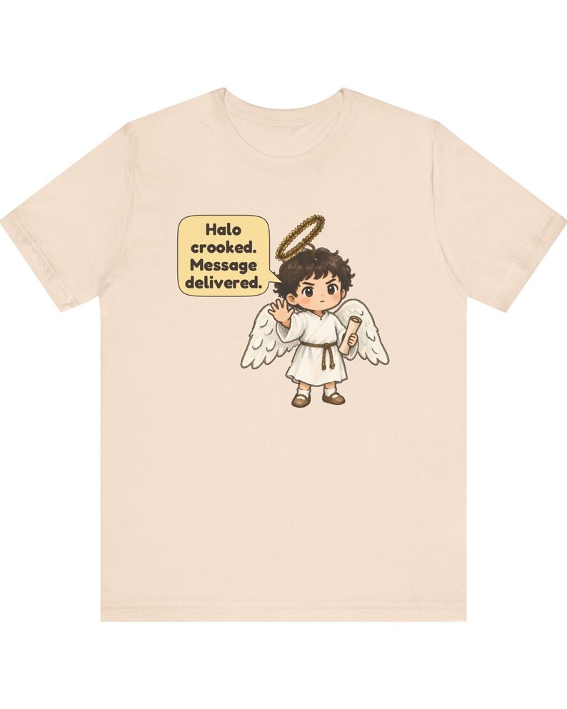 May include: A light beige t-shirt featuring a graphic of a cartoon angel with dark hair, white wings, and a crooked halo. The angel holds a scroll and waves, with a speech bubble containing the text: Halo crooked. Message delivered.