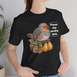 K&ouml;nnte beinhalten: Ein schwarzes T-Shirt mit einer Grafik eines runden grauen Vogels mit orangefarbenem Gesicht, der auf einem Ast mit zwei gelben Birnen und zwei kleinen Geschenkkartons sitzt. Eine Sprechblase neben dem Vogel enth&auml;lt den Text, Please stop sending birds.