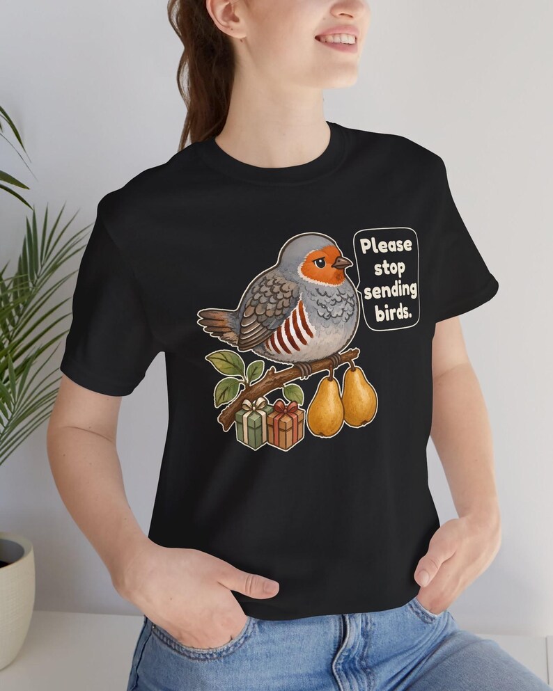 K&ouml;nnte beinhalten: Ein schwarzes T-Shirt mit einer Grafik eines runden grauen Vogels mit orangefarbenem Gesicht, der auf einem Ast mit zwei gelben Birnen und zwei kleinen Geschenkkartons sitzt. Eine Sprechblase neben dem Vogel enth&auml;lt den Text, Please stop sending birds.