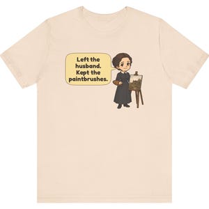 May include: A light beige t-shirt featuring a cartoon illustration of a person in a dark dress standing next to an easel with a painting. A speech bubble contains the text: Left the husband. Kept the paintbrushes.