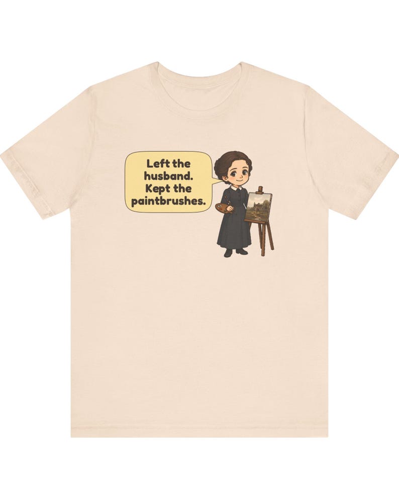 May include: A light beige t-shirt featuring a cartoon illustration of a person in a dark dress standing next to an easel with a painting. A speech bubble contains the text: Left the husband. Kept the paintbrushes.